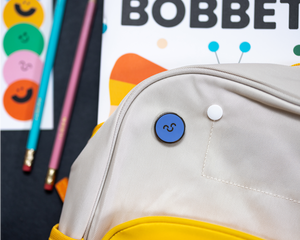 Blue pin with happy face on a kid's backpack sitting on top of a Bobbety magazine, bookmark, and pencils.