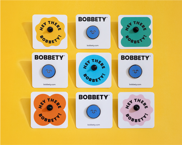 Nine Bobbety Blue Pins attached pin backers sitting in a grid on a yellow background