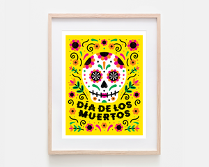 A framed art print featuring a colorful Dia de los Muertos sugar skull design on a bright yellow background, housed in a light natural wood frame with a white mat.