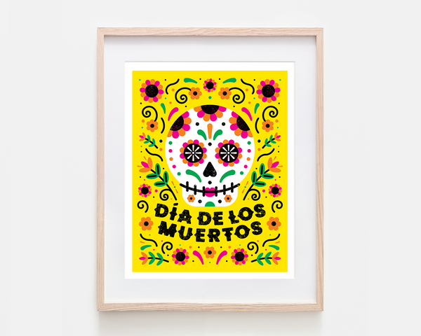 A framed art print featuring a colorful Dia de los Muertos sugar skull design on a bright yellow background, housed in a light natural wood frame with a white mat.
