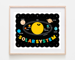 A framed children's educational poster of the Solar System, featuring a cute cartoon sun, planets, and the word 'SOLARSYSTEM' in colorful letters on a black background, housed in a light wood frame.