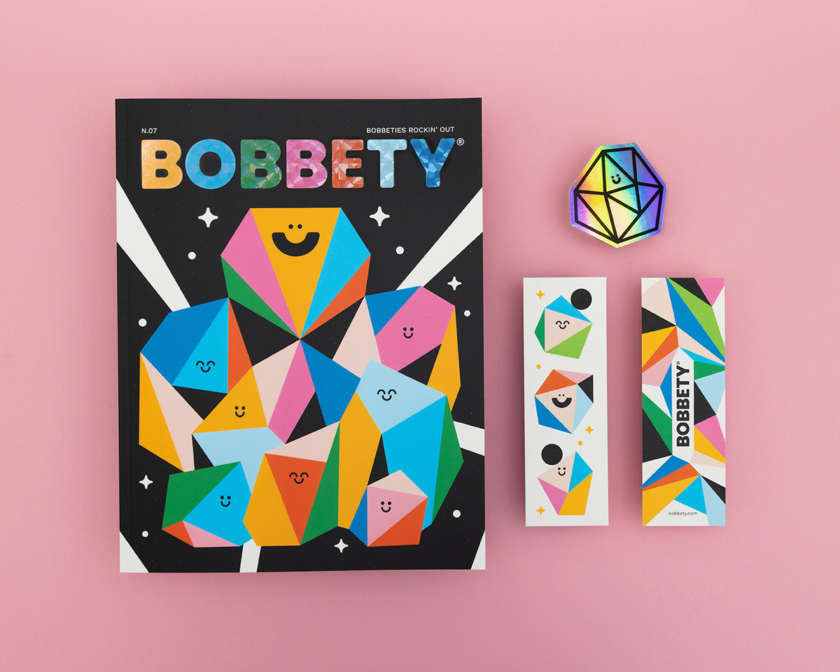 A flat-lay product shot of the Bobbety book and its accessories on a solid pink background. The set includes a black hardcover book with a colorful geometric cover, two matching bookmarks, and a small colorful polyhedron sticker