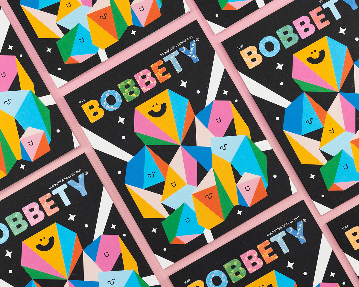 A repeating flat-lay pattern of colorful children's book covers titled BOBBETY'. Each cover has a black background with a large multicolored geometric character made of smiling triangles and the word 'BOBBETY' in large, playful letters.